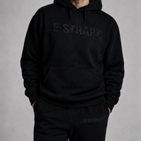 A man wearing a black E-STRAPZ hoodie and matching black sweatpants, standing against a neutral gray studio background.