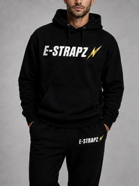 A man wearing a black pullover hoodie and matching sweatpants with the brand name E-STRAPZ printed in white with a gold lightning bolt on the chest and thigh, set against a neutral gray studio background.